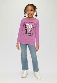 Paarse langemouwen shirt met metallic "CUTE" tekst, gecombineerd met lichtblauwe spijkerbroek en witte sneakers. Gladde stof, ontspannen pasvorm.