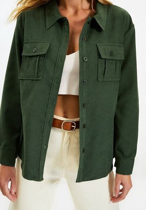 Vrouw die een donkergroen overhemd met klepzakken draagt over een wit cropped topje, crème broek en een bruine riem met een gouden gesp.