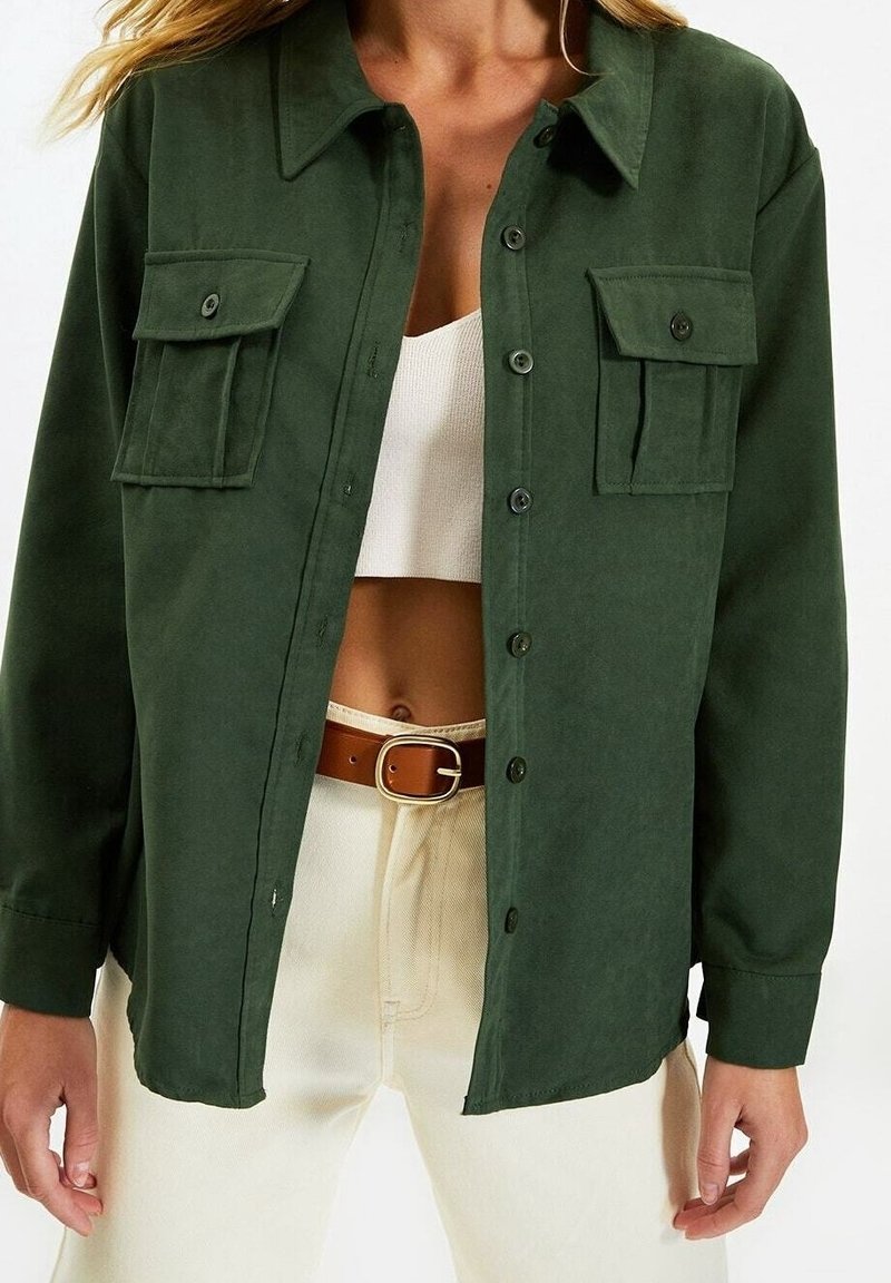 Femme portant une chemise boutonnée vert foncé avec poches à rabat sur un crop top blanc, pantalon crème et ceinture marron avec boucle dorée.