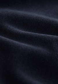 Tamsiai mėlyna audinio medžiaga su minkštu, tekstūruotu paviršiumi ir subtiliais įstrižais pynimo raštais, sukabinta taip, kad atskleistų jos sklandžias, tekančias savybes.