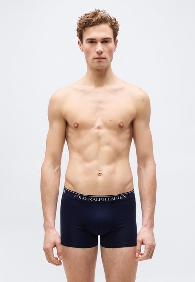 Νεαρός άνδρας με σγουρά μαλλιά που φοράει σκούρο μπλε εσώρουχα Polo Ralph Lauren, στέκεται μπροστά σε ένα απλό ανοιχτόχρωμο φόντο.