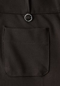 Un tissu noir avec une texture lisse présente une poche carrée cousue sur le côté droit et un accent en matériel circulaire argenté au-dessus.