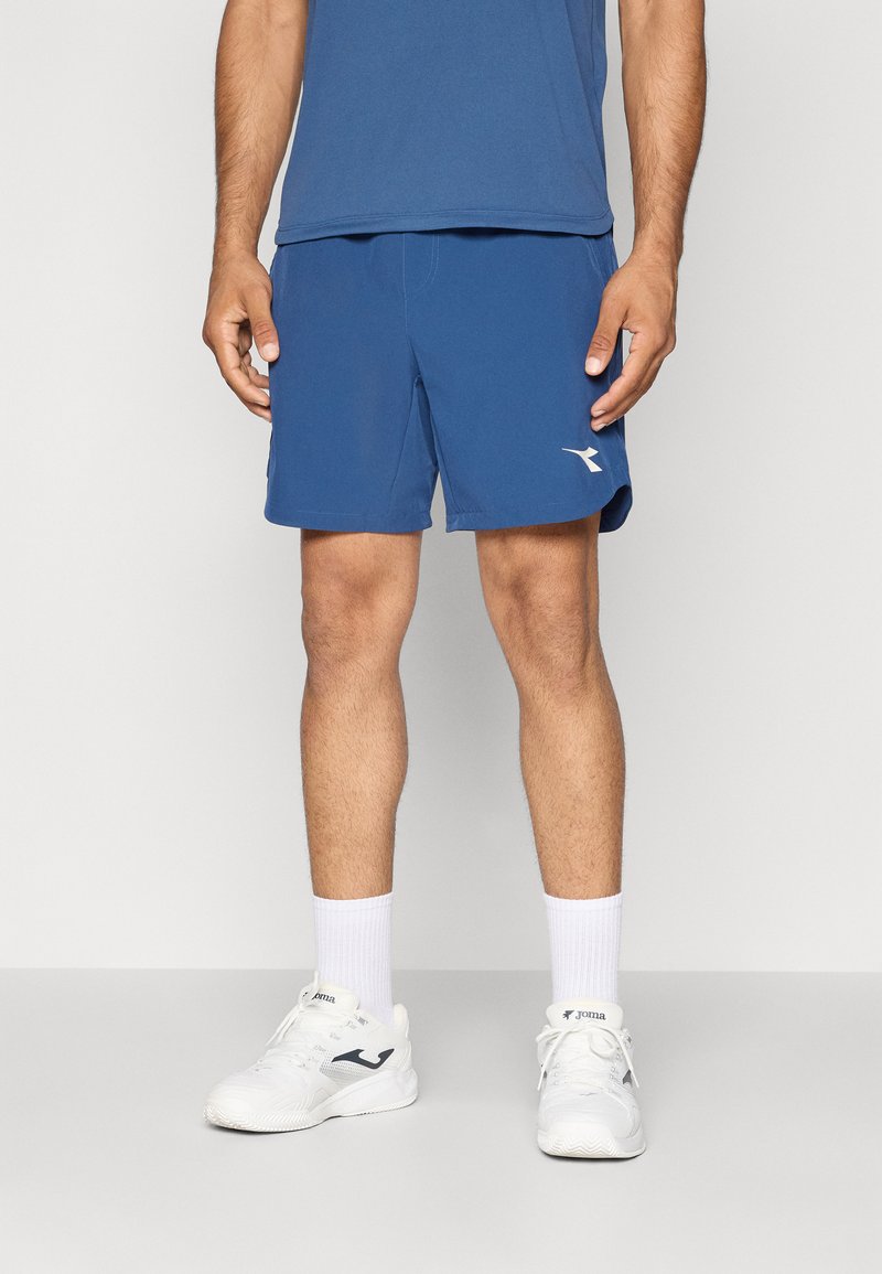 Blaue Sportshorts aus leichtem Material, mit elastischem Bund und einem weißen Logo auf der linken Seite. Getragen mit weißen Sneakers und Socken.