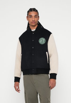 Chaqueta varsity negra y crema con cierre de botones, puños acanalados y un logo bordado verde redondo en el pecho. Tejido texturizado.