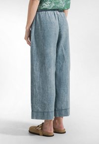 Lichtblauwe wijde broek van linnen met een elastische tailleband en opgerolde pijpen. Draagt daarbij bruine instapschoenen, waardoor de gestructureerde stof zichtbaar is.