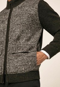 Uomo che indossa un maglione con zip, maniche scure e pannello frontale grigio a scacchi, con la mano in tasca, su uno sfondo chiaro.