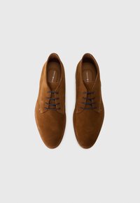 Chaussures habillées pour hommes en daim marron, à lacets foncés, présentées côte à côte sur un fond gris clair uni.