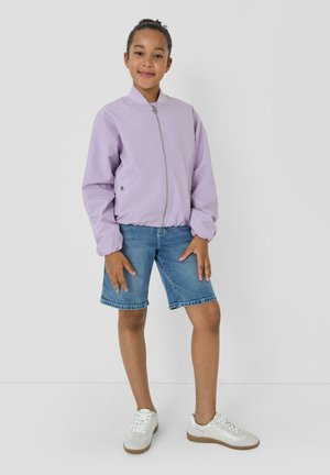 Jonge persoon staand, gekleed in een lichtpaarse jas met ritssluiting, denim shorts tot de knieën en witte sneakers, tegen een effen achtergrond.