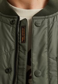 Giacca bomber trapuntata verde con colletto a costine, chiusura con zip e dettagli con bottoni a pressione. Presenta un'etichetta del logo sul davanti. La fodera interna è leggera.