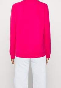 Fuchsiová mikina s kulatým výstřihem a dlouhými rukávy, s manžetami a pasem, v kombinaci se světlými denimovými kalhotami. Hladká textura, jednobarevný design.