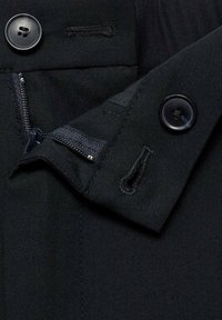 Gros plan sur un tissu noir avec une fermeture éclair partiellement dézippée et deux boutons noirs avec des boutonnières sur des rabats superposés.