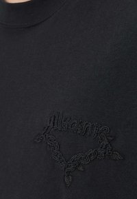 Sort bomulds t-shirt med et tekstureret sort broderet motiv og rund halsudskæring. Stoffet fremstår glat med en let glans.