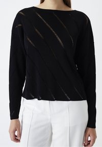 Czarny longsleeve z diagonalnymi wycięciami i fakturowanym materiałem, w połączeniu z białymi spodniami w szerokim kroju. Prosty, luźny fason.