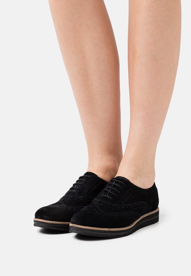 Paire de chaussures brogues en daim noir à lacets portées pieds nus, vues de face latérale avec des coutures détaillées et un bord marron clair.