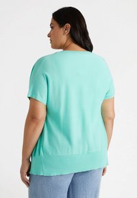 Turkos kortärmad topp med avslappnad passform, mjuk textur och en sänkt nederkant bak, kombinerad med ljusblå jeans.