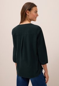 Blusa verde scuro a maniche lunghe con scollo rotondo e dettagli di pieghe sul retro. Tessuto morbido e vestibilità comoda, abbinata a jeans di denim blu.