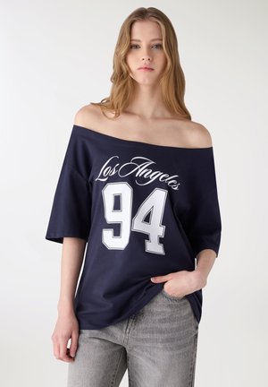 Giovane donna che indossa una maglietta blu navy con la scritta "Los Angeles 94" e jeans grigi chiari, in piedi con una mano in tasca.