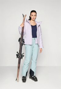 Lichtpaarse gewatteerde ski-jas, blauw shirt, lichtblauwe skibroek, zwarte skischoenen, een snowboard met donkere bindingen vasthoudend.