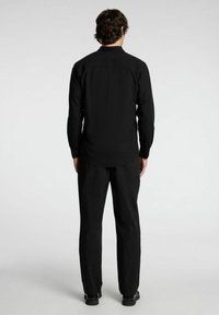 Camicia nera a maniche lunghe con polsini bottone, texture liscia e orlo arrotondato, indossata con pantaloni neri abbinati, vista da dietro.