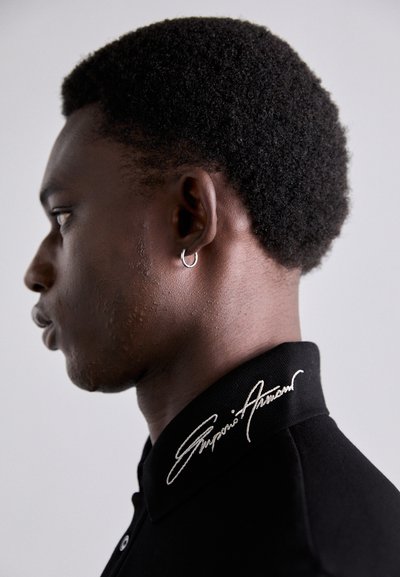 Svart polo skjorta med hög krage som har vit broderad text på kragen. Nära profilbild av en modell med mörkt lockigt hår.