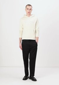 Sudadera crema con logo "GUESS" en relieve, combinada con pantalones negros y zapatos negros. Textura suave; diseño con capucha; corte clásico.