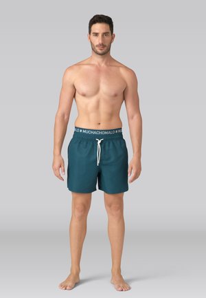 Muž stojaci naboso, oblečený v tyrkysových plavkových šortkách s pásom "MUCHACHOMALO" a bielou šnúrkou, na pozadí jednoduchého šedého pozadia.