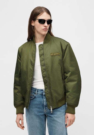 Άτομο που φοράει μαύρα γυαλιά ηλίου, χακί bomber μπουφάν με κεντημένη ετικέτα "Off-White", λευκό πουκάμισο και μπλε τζιν, μπροστά σε απλό φόντο.