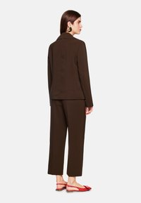 Brauner maßgeschneiderter Anzug mit langen Ärmeln, weit geschnittenen Hosen und roten flachen Sandalen, der ein minimalistisches Design und eine glatte Stoffstruktur aufweist.