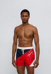 Μαγιό κόκκινου και ναυτικού μπλε χρώματος με διαγώνιο σχέδιο, λευκό κορδόνι και το λογότυπο «Tommy». Κατασκευασμένο από ελαφρύ ύφασμα με λεία υφή.