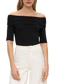 Donna che indossa un top nero senza spalle con maniche lunghe fino ai gomiti, abbinato a pantaloni bianchi a vita alta, in piedi davanti a uno sfondo bianco.