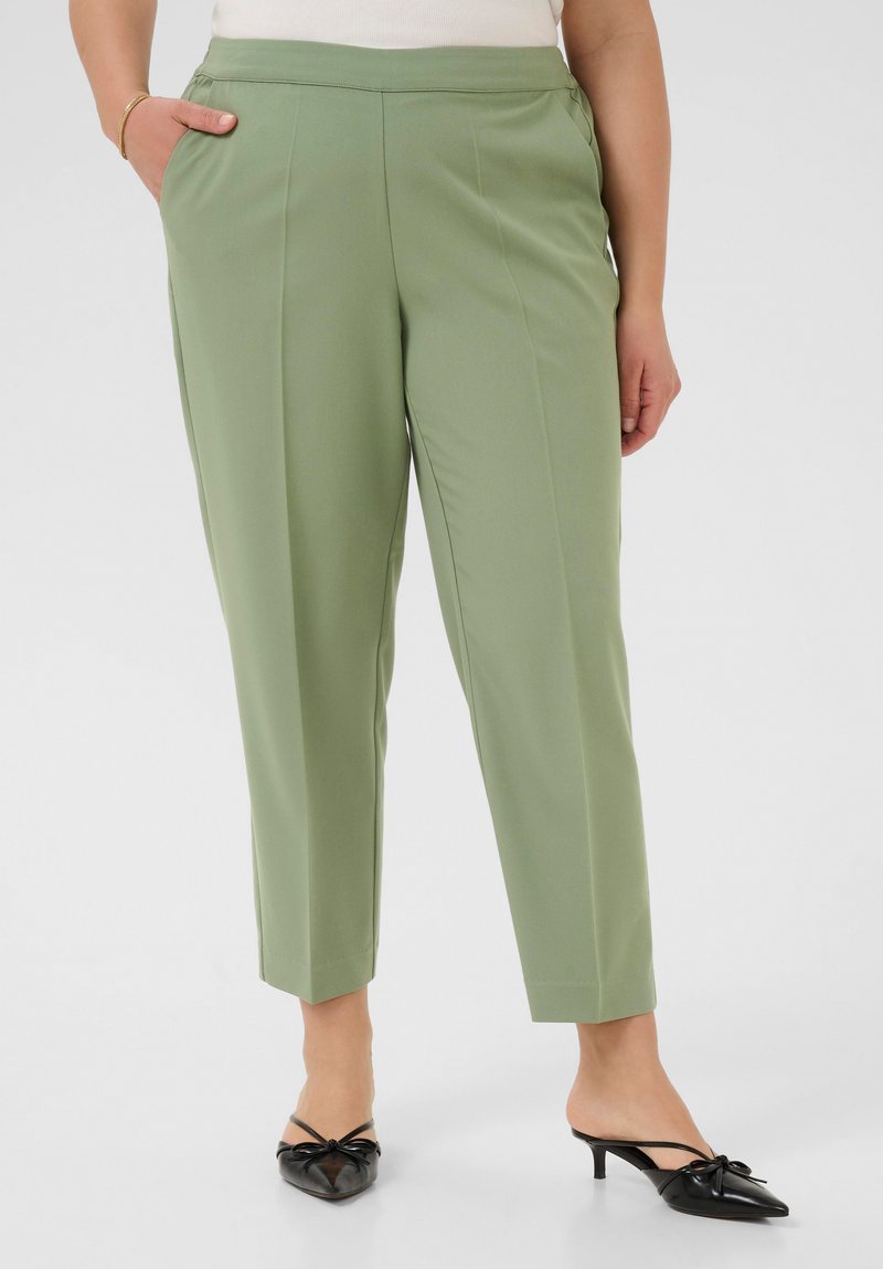 Pantaloni verdi chiaro su misura, caratterizzati da un tessuto liscio, vestibilità slim, due tasche laterali e pieghe discrete. Abbinati a sandali neri con cinturino posteriore.