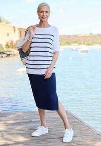 Gestreifter weißer und marineblauer Strickpullover mit rundem Ausschnitt, kombiniert mit einem marineblauen Bleistiftrock und weißen Sneakers. Hält eine geflochtene Tragetasche.