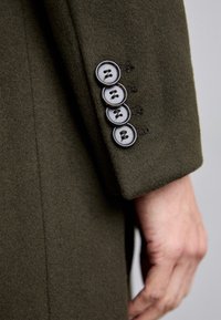 Manica di un cappotto di lana verde oliva con quattro bottoni scuri, caratterizzato da cuciture decorative e una finitura morbida e testurizzata.
