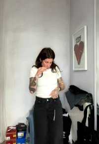 Giovane donna con tatuaggi sistema la sua camicia bianca in una stanza con scatole di scarpe impilate, vestiti sul termosifone e un quadro incorniciato a forma di cuore sulla parete.