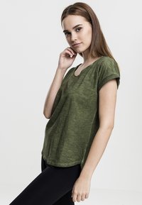 T-shirt verde a maniche corte con colletto rotondo e orlo curvo, realizzato in tessuto strutturato. Modello abbinato a leggings neri, in posizione di profilo.