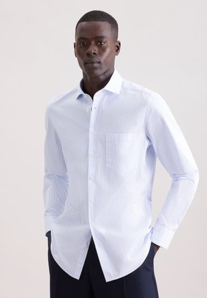 Mężczyzna w jasno-niebiesko-białej koszuli w paski z długim rękawem i ciemnych spodniach, stojący na jednolitym tle.