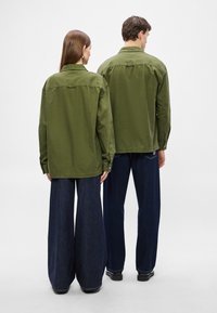 Πράσινα βαμβακερά πουκάμισα πάνω σε δύο φιγούρες, εμφανιζόμενα από πίσω. Χαλαρή εφαρμογή με μανσέτες με κουμπιά και ορατό πίσω καβάλο; συνδυασμένα με σκούρα τζιν φαρδιάς γραμμής.