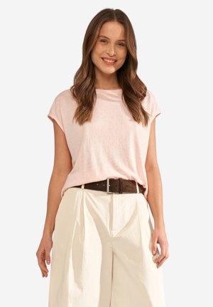 Žena s dlouhými hnědými vlasy, která má na sobě světle růžový top bez rukávů, krémové kalhoty s vysokým pasem a tmavě hnědý pásek, usmívá se do kamery.