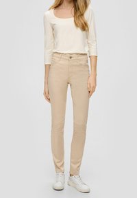 Jeans ajustados beige con cinco bolsillos, un cierre de cremallera y un botón. Combinados con una camisa blanca de manga larga y zapatillas blancas.