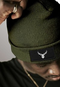 Khaki beanie with vertical ribbed texture; features a black rectangular patch with a white deer logo. Worn slightly tilted.