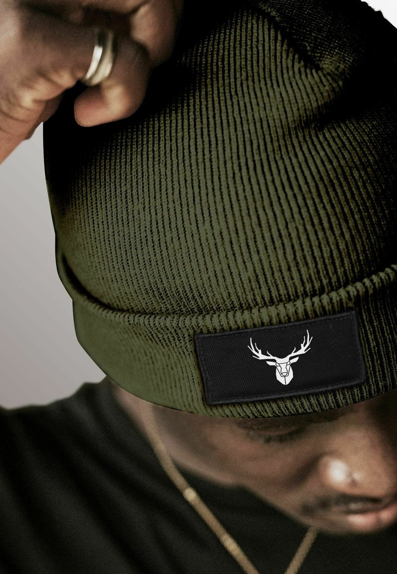 Khaki beanie with vertical ribbed texture; features a black rectangular patch with a white deer logo. Worn slightly tilted.