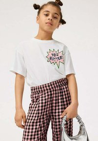 Hvid t-shirt med kortærmer og grafikken "DU BETYDER NOGET!" i pink og grøn. Sammen med sorte og pink ternede bukser, holder en sølvfarvet taske.