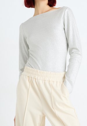 Femme portant un haut argenté brillant à manches longues, rentré dans un pantalon crème taille haute avec ceinture élastique, les mains dans les poches.