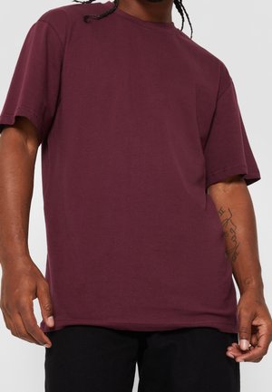 Mężczyzna z zaplecionymi włosami nosi gładki, bordowy t-shirt z krótkim rękawem i czarne spodnie, pokazując tatuaże na przedramionach na jednolitym tle.