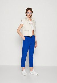 Béžová vatovaná vesta s vysokým límcem, modré kalhoty na míru a bílé tenisky; modelka má také průhledné sluneční brýle a krátký bílý top.