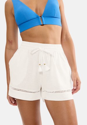 Vrouw met een gestructureerde blauwe crop top en witte shorts met trekkoord en kanten rand, handen ontspannen langs het lichaam, vlindertattoo op de linkerarm.