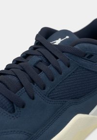 Κοντινός πλάνο ενός ναυτικού μπλε αθλητικού παπουτσιού με υφή στο πλάι, χοντρά κορδόνια, επενδυμένο κολάρο και λευκό λογότυπο στη γλώσσα πάνω από τη λευκή σόλα.