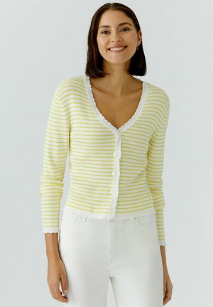 Femme souriante portant un cardigan rayé blanc et jaune clair boutonné, avec un pantalon blanc, debout devant un fond clair uni.