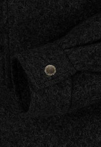 Black wool fabric with a textured surface, featuring a gold metallic button on the cuff, which has an embossed design around its edge.