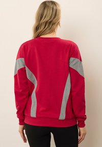 Donna con capelli biondi lunghi che indossa un felpa rossa con pannelli geometrici grigi sul retro e pantaloni neri, in piedi contro un muro semplice.